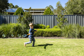 Image of young boy running, playing with bubbles with a grey colorbond fence in the back